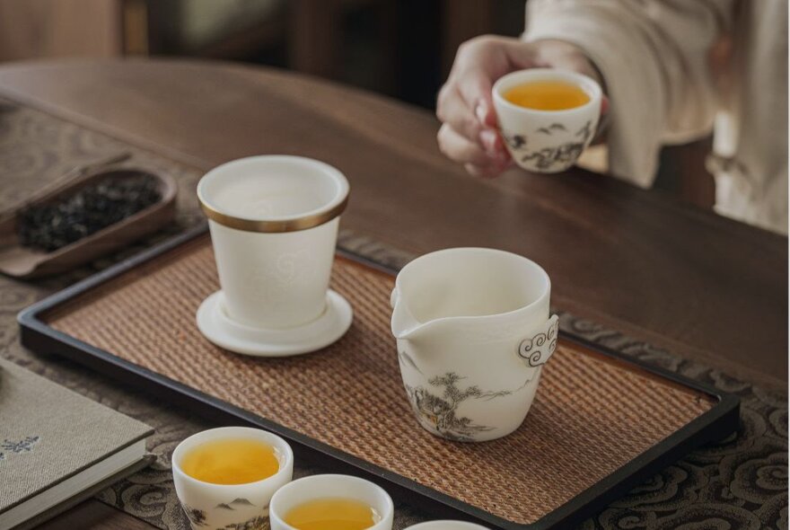 A hand holding a small cup of tea, in the foreground are more small cups of tea and a ceramic pot. 