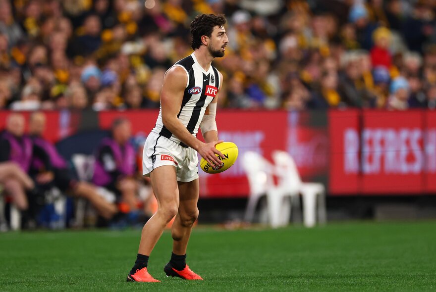 Collingwood player Josh Daicos looks across the ground while holding a yellow football, a blurred crowd and two white plastic chairs in the background.