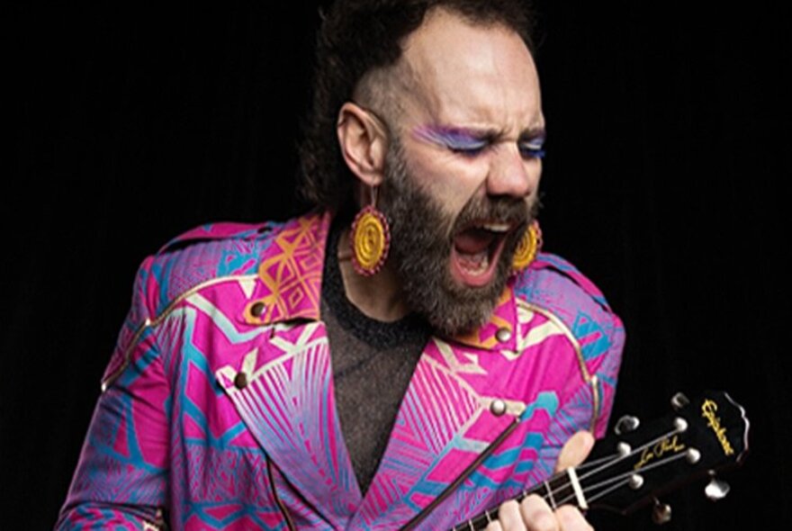 A man with a beard, pink and purple eye makeup, large gold earrings and a brightly patterned pink and purple jacket, playing guitar and singing.