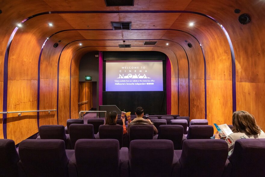 Three people sitting in a movie theatre with purple seats and wooden walls.