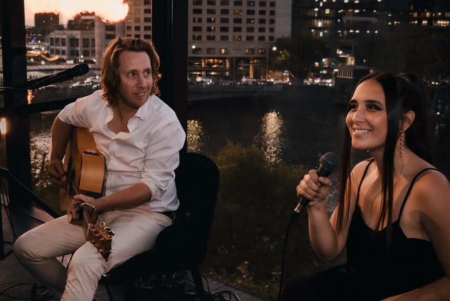 A man playing guitar while a woman holds a microphone and sings, with a view of the Yarra river in the background.