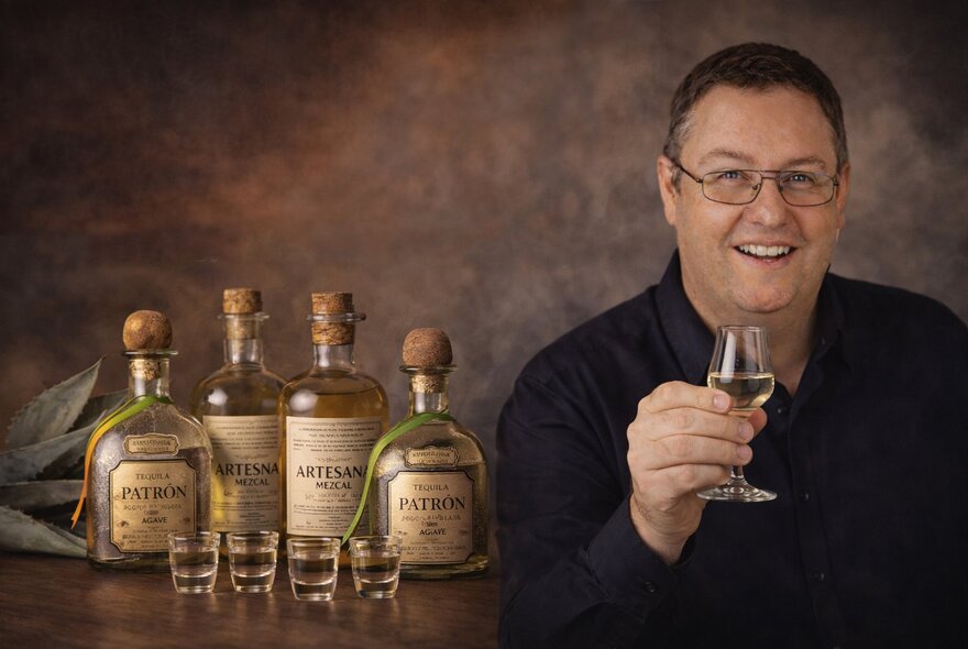 A smiling man holding a glass of tequila, bottles and shot glasses beside him. 