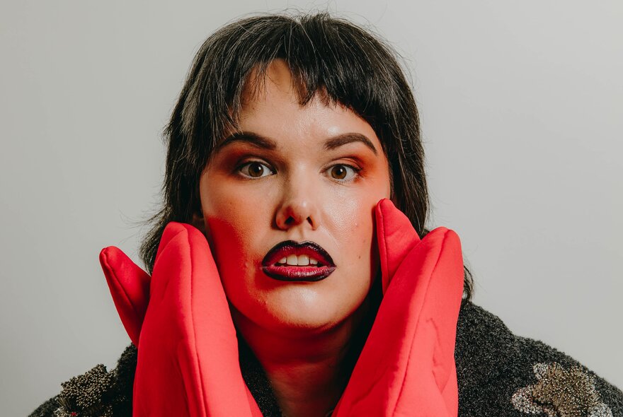 A close-up portrait of a person with dark hair, a short fringe, looking straight ahead with an open-mouthed expression, framing their face with both hands while wearing bright red, quilted oven mitts.