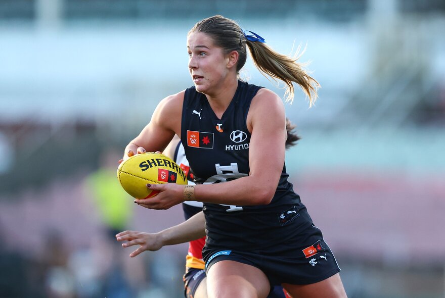 A Carlton AFLW player takes off with the football, ponytail flying out the back. 