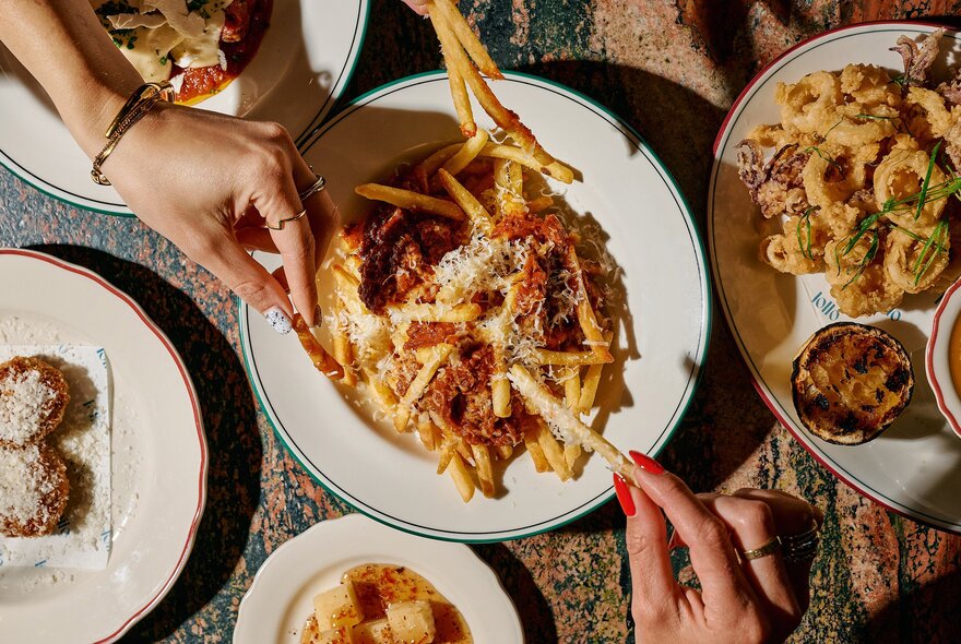 Hands reaching for topped fries from a range of dishes on a restaurant table.