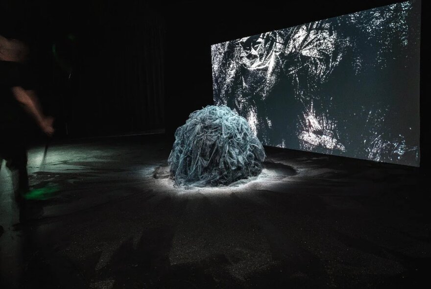 A large, mounded form made from strips of fabric, spotlit in front of a large screen in an exhibition space.