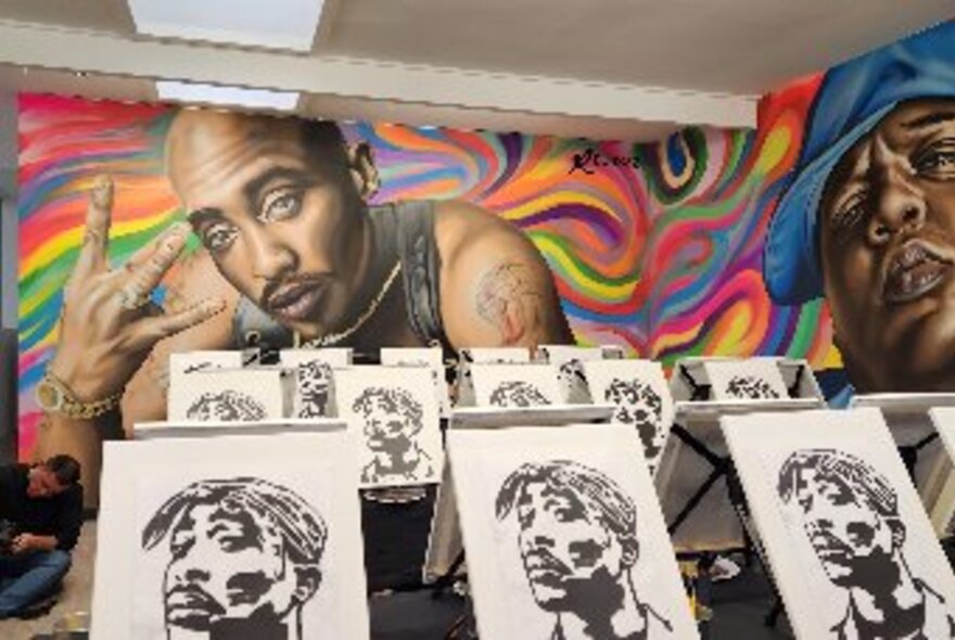 Several rows of canvases in a room with large, wall murals of two hip-hop styled men against colourful swirls, at rear and to right.