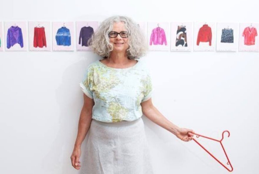 A woman holding a coat hanger in front of a wall with small paper clothing cut-outs arranged in a line. 