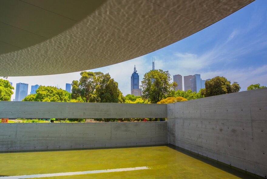 A concrete pavilion with a pond. There is a city skyline in the background. 