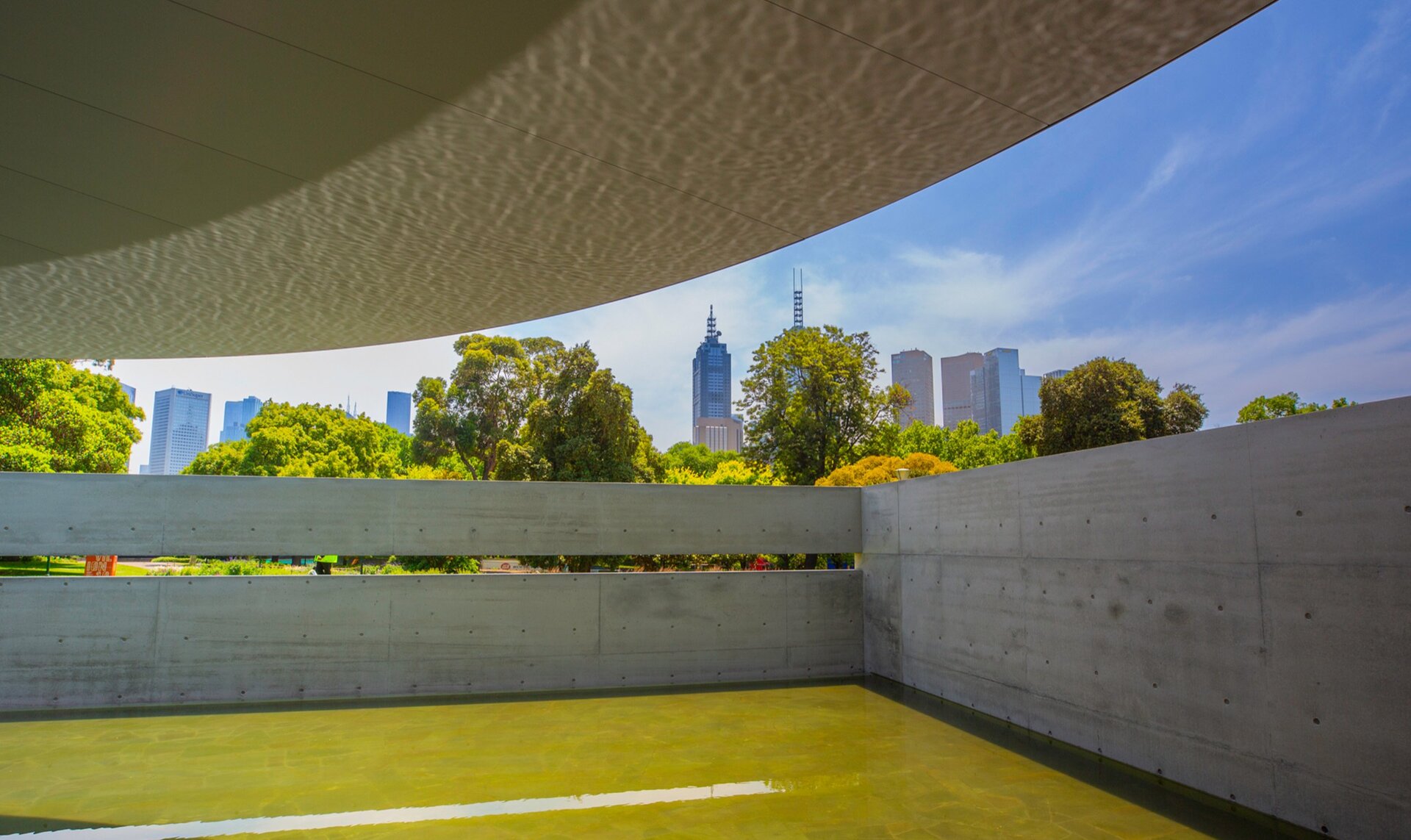 A concrete pavilion with a pond. There is a city skyline in the background. 