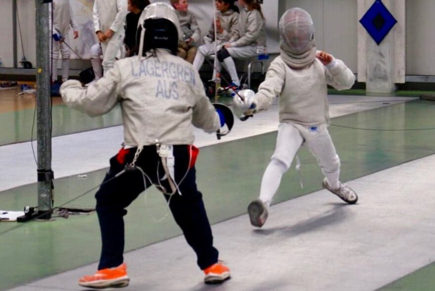 Two children dressed in white fencing outfits, fitted with head guards, fencing with swords on a fencing mat, indoors.