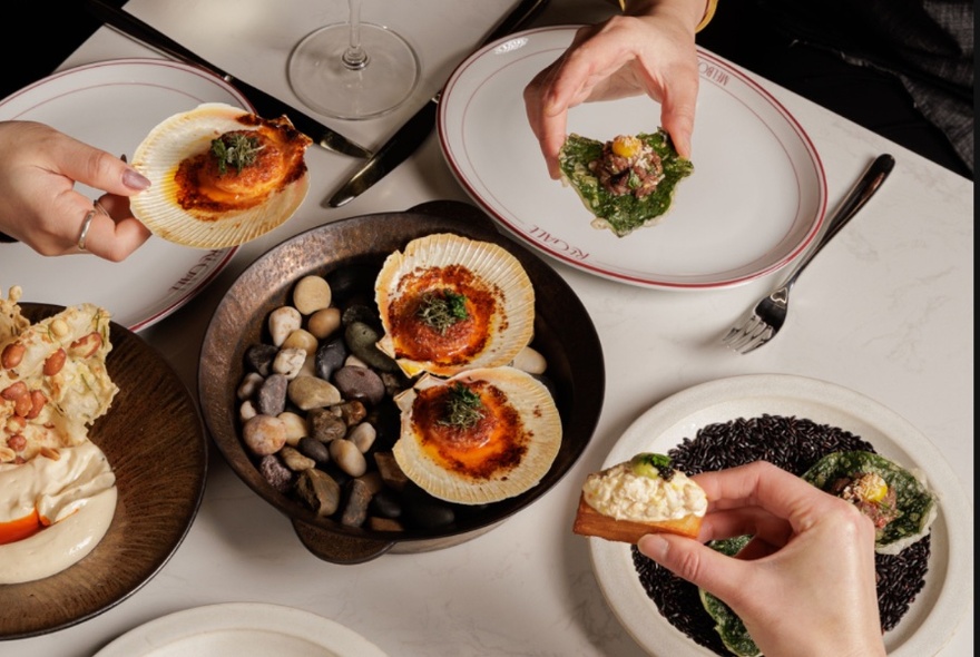 A selection of seafood dishes seen from above, with three hands each holding a bite-sized portion. 