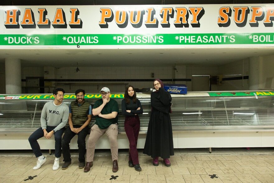 Five people standing in front of an empty poultry stall at a market, with signage above them.