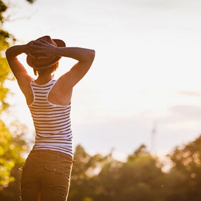 The back of a woman arching her back with her hands on her head looking at the sky with a sunny glow all around her. 