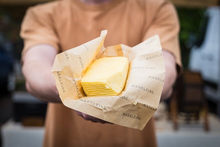 Close up of a knob of butter in wax paper wrapping, being held by a man in an orange t-shirt. 