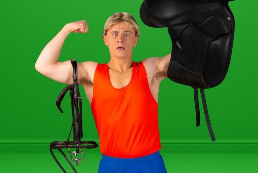 Young man with blond wig, orange singlet and tracksuit pants holding riding equipment.