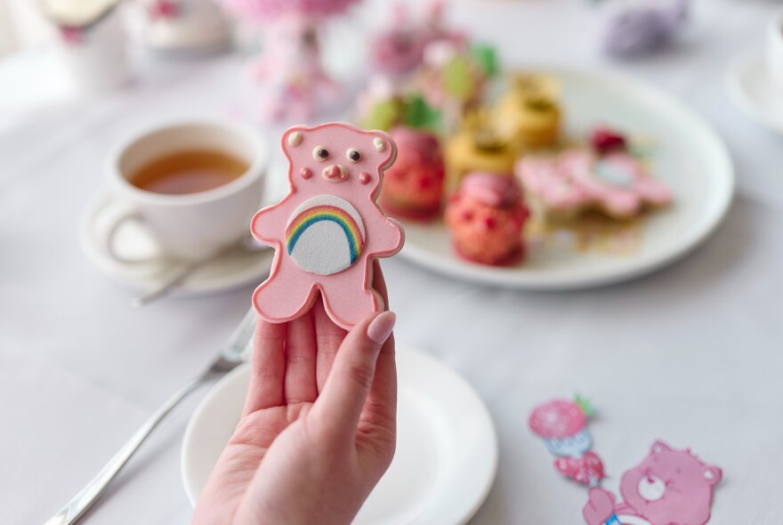 A woman's hand holding a Care Bear-shaped cookie with pink icing and a rainbow on its tummy; other iced treats blurred in the background.