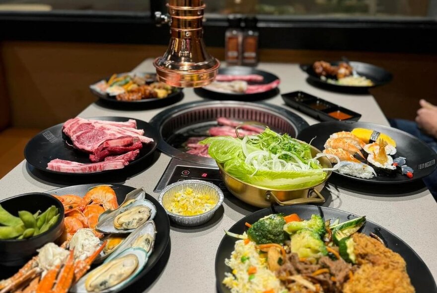 Table at a Korean BBQ restaurant with a smokeless grill in the middle and multiple plates of Korean food including fried chicken, salad, fried rice, crab, mussels, prawns, edamame, corn cheese, gem lettuce, white onion, sushi, raw meat and dipping sauces.