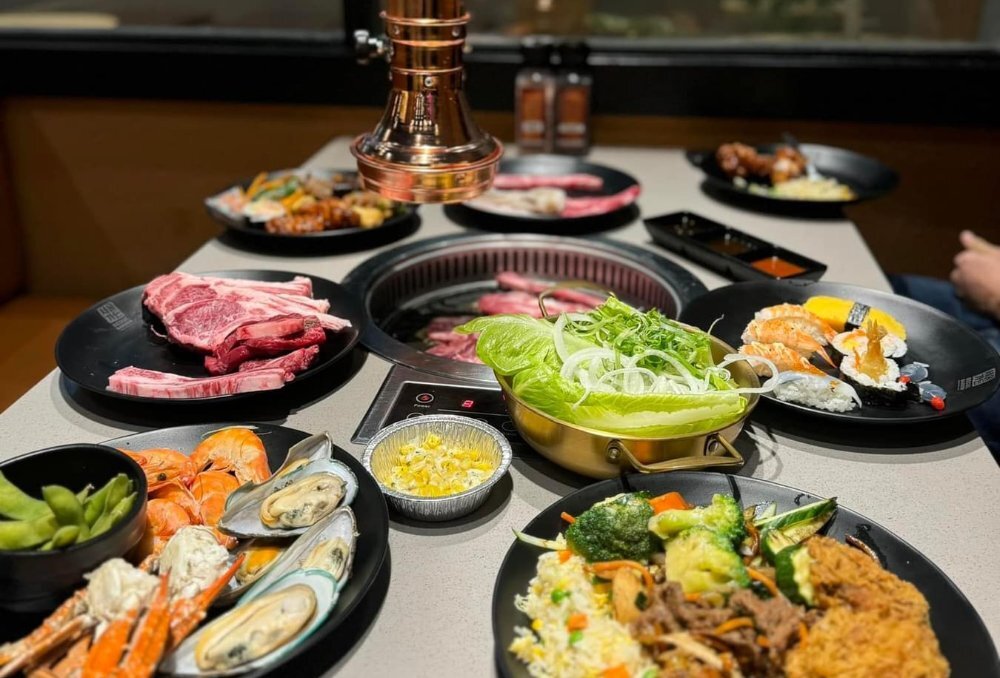 Table at a Korean BBQ restaurant with a smokeless grill in the middle and multiple plates of Korean food including fried chicken, salad, fried rice, crab, mussels, prawns, edamame, corn cheese, gem lettuce, white onion, sushi, raw meat and dipping sauces.