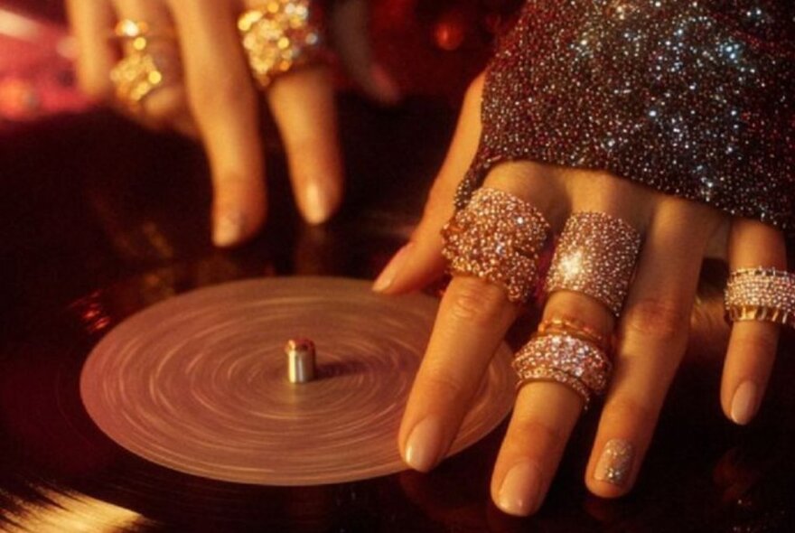 Hands with many diamanté rings on fingers handling a vinyl record. 