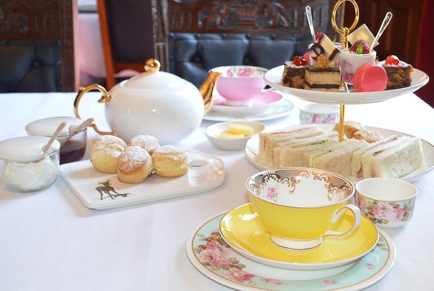 Afternoon tea setting with scones, cake stand with sandwiches and cakes, teapot, cups and saucers.