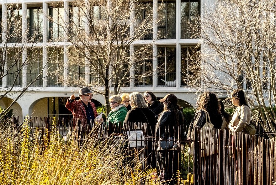 A small group of people on a tour on a pathway with yellow grass on one side and a large building on the other.
