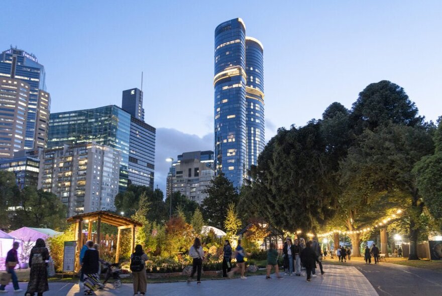 People enjoying a twilight session at Melbourne Flowers & Garden Show in the illuminated Carlton Gardens with the Melbourne city skyline, including the distinctive tall buildings, in the background.