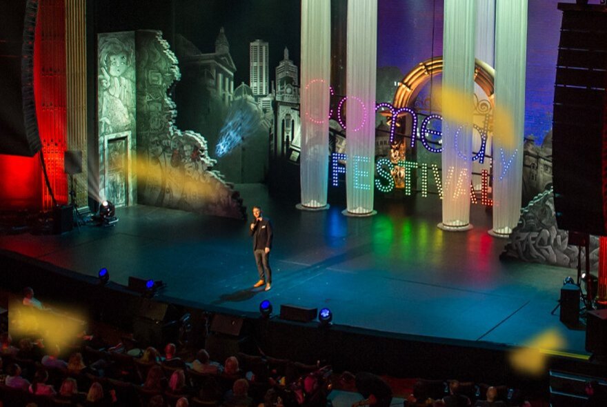 A lone comedian on a huge stage in front of an audience, the backdrop is colourful and has the word FESTIVAL across an image of Melbourne. 