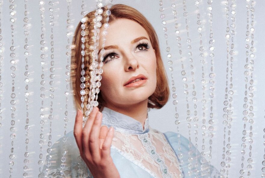 Comedian, Abby Howells, wearing a prim, blue lace blouse, holding a crystal hanging curtain away from her peaceful face.