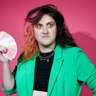Comedian and musician Tricksy Collins, wearing a vibrant green jacket and holding a fanned out deck of cards.
