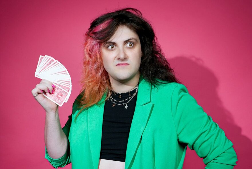 Comedian and musician Tricksy Collins, wearing a vibrant green jacket and holding a fanned out deck of cards.