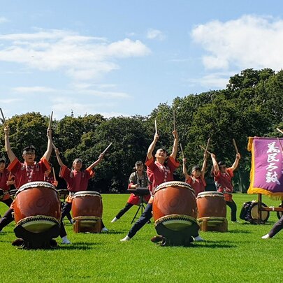 Gimin Taiko drumming performers outdoors in a park setting with their large drums and hands in the air. 