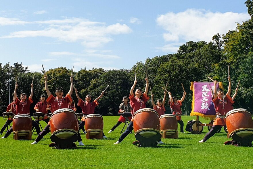 Gimin Taiko drumming performers outdoors in a park setting with their large drums and hands in the air. 