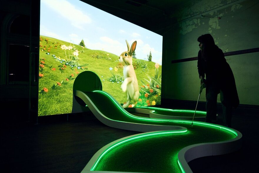 A curving mini-golf putting green, highlighted in green neon coming out of an arched hole in a projector screen that shows a bunny standing on its hind legs in a grassy meadow with wildflowers.