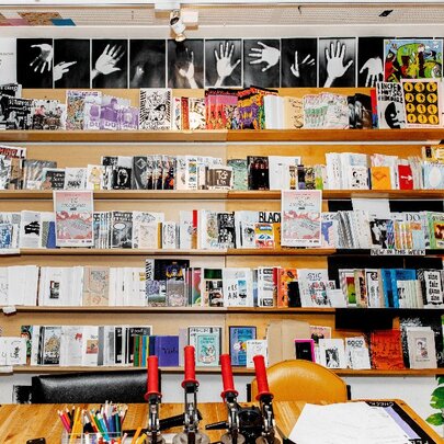 The studio space at Sticky Institute, with a wall of shelves against the rear wall, and a large work bench with paper, and tools for making zines.