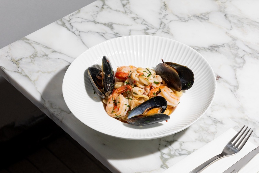 An Italian marinara dish with three muscles and prawns, on a marble table with a fork and knife on a white napkin.