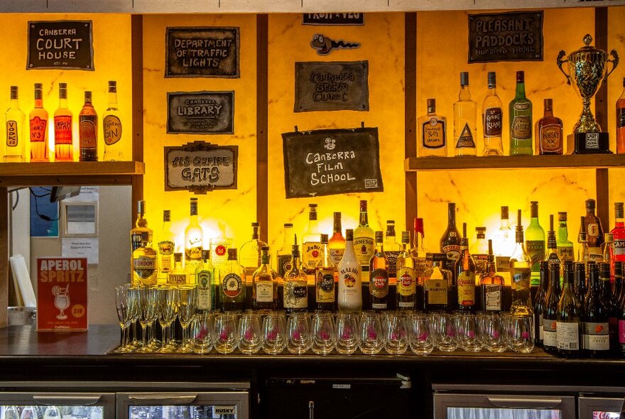 Bar area with rows of bottles against a backlit glass wall with signs.