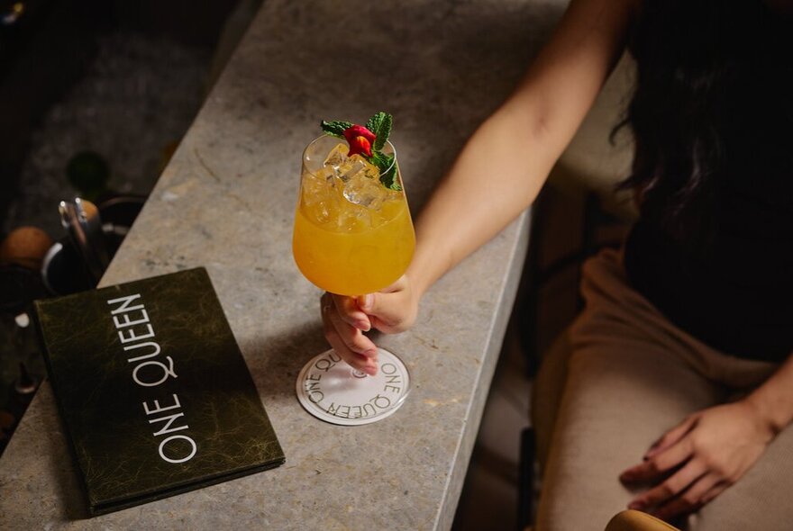 A person's arm resting on a bar, their hand holding the stem of a cocktail glass, a drinks menu in front of the glass.