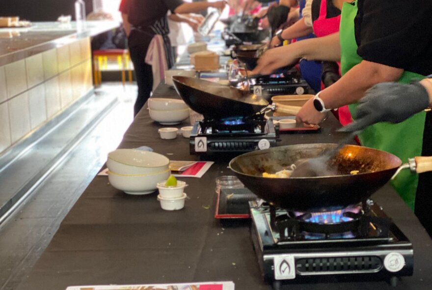 Long row of people preparing Asian food on portable gas stoves on a long counter.