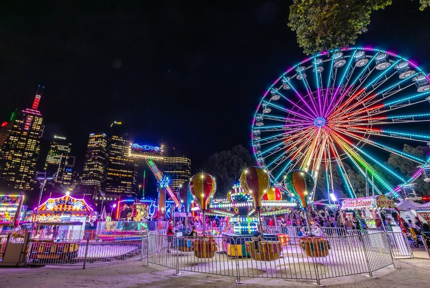 A Ferris wheel and rides lit up in colourful lights at a city carnival at night.