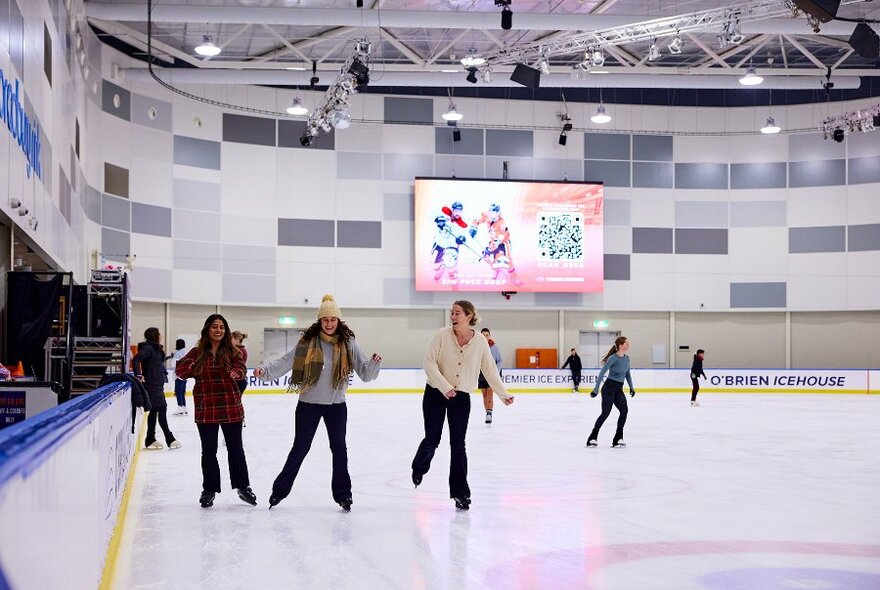 Three friends ice skating on an indoor rink.