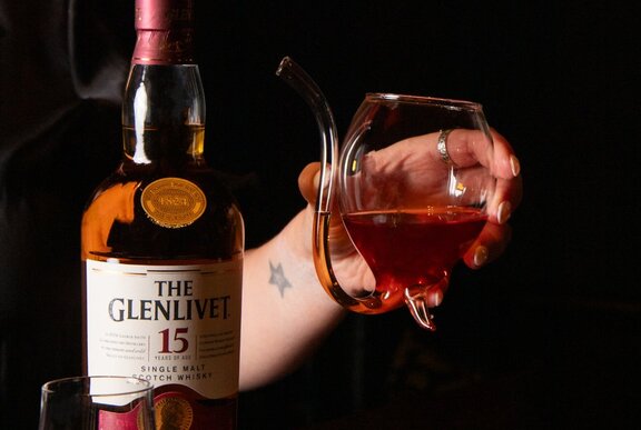 A bottle of Glenlivet Whisky next to a hand holding a cocktail in a unique glass with a curved stem from the bottom that functions as a straw.