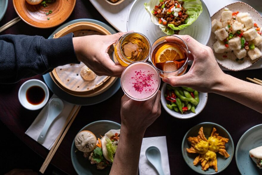 Three hands clinking beverages in glasses, viewed from above, with plates of food resting on the table underneath.