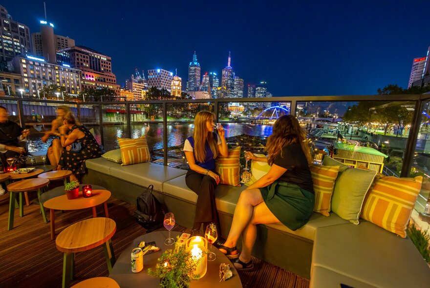 Two friends are drinking wine at a riverside rooftop bar with the city skyline in the background