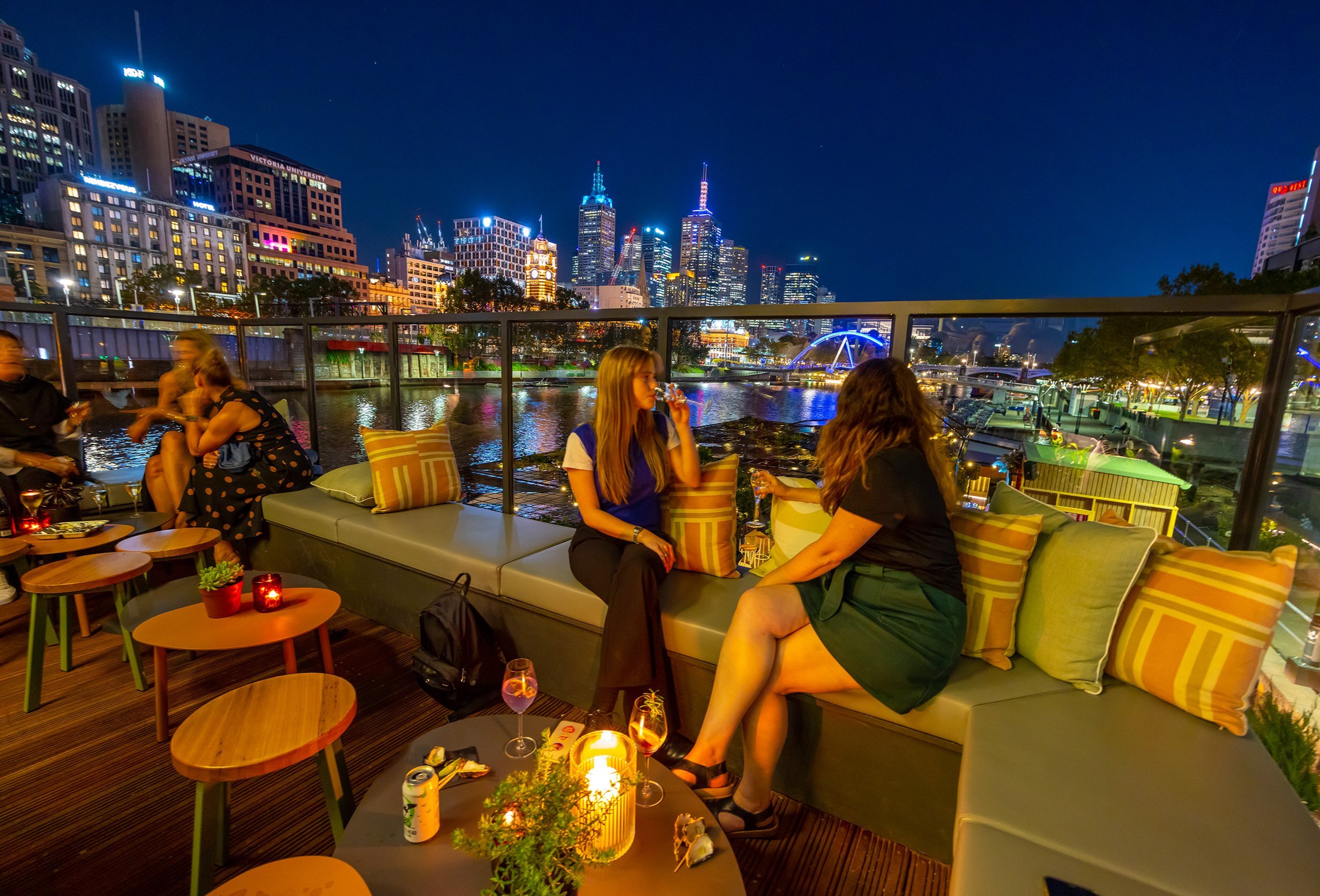 Two friends are drinking wine at a riverside rooftop bar with the city skyline in the background