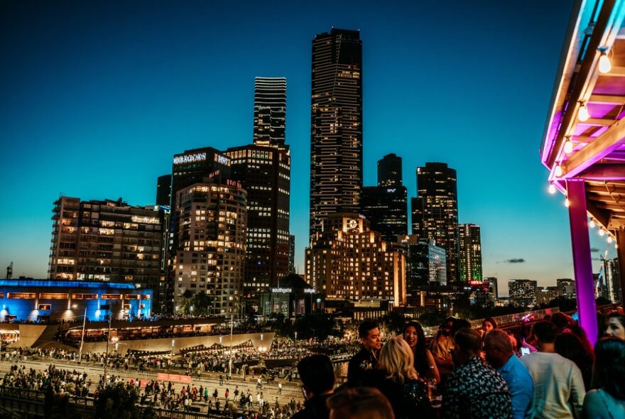 A twilight view of the Melbourne City Skyline with a rooftop bar visible in the bottom right corner, packed with people. 