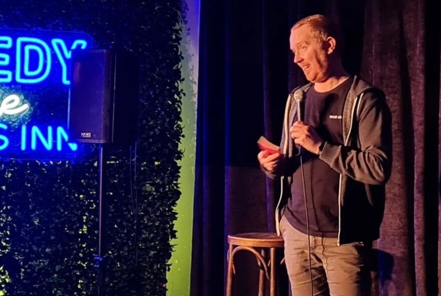 Male stand-up comedian talking into a microphone and standing on a stage, partial neon signage visible on the left of the image.