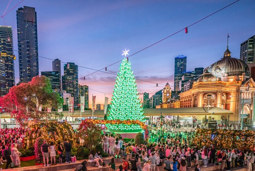 A giant illuminated Christmas Tree at Federation Square, surrounded by people and view of Flinders St Station and city buildings in the background. 