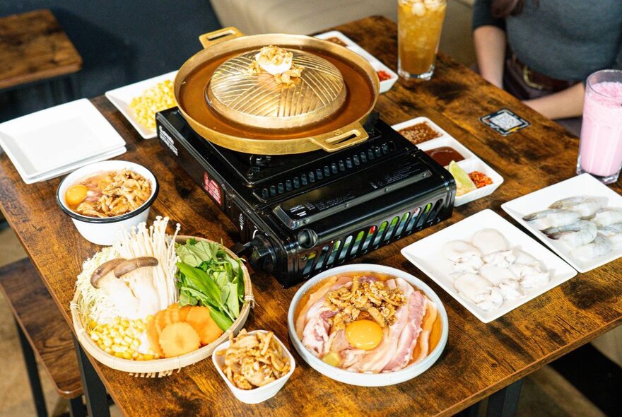 Dishes of Asian food surrounding a central cooking stove topped with a lidded wok on a restaurant table.