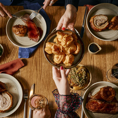 Someone passing a plate of Yorkshire puddings over a table set with rolled pork roast, veggies, wine and gravy.